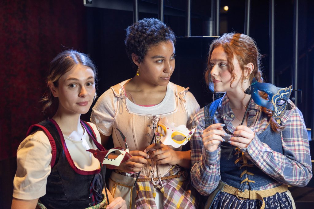 Three feminine people in mixed contemporary and renaissance clothing stand before the camera. The person on the left looks directly towards the viewer, while the other two look at each other, seeming to be in conversation.
